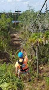 A man glides on a zipline at Empower Adventures Tampa Bay zip-line park through a narrow pass of palm trees.